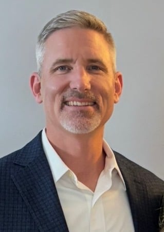 Professional headshot of a smiling man with gray hair wearing a dark blazer and white dress shirt against a neutral background